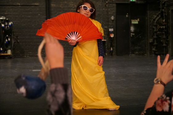 Foto: Ein Kind steht mit großer Sonnenbrille, langem Wickelkleid und großem Fächer vor Publikum.