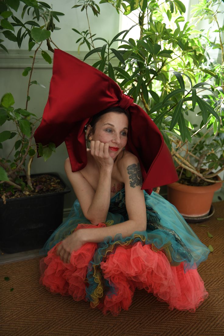 Meret Becker Foto: Meret Becker sitzt in einem Kleid aus türkis-und leuchten rosa-farbenem Kleid mit einer großen roten Schleife auf dem Kopf in der Hocke. Der Hintergrund zeigt verschiedene Zimmerpflanzen.
