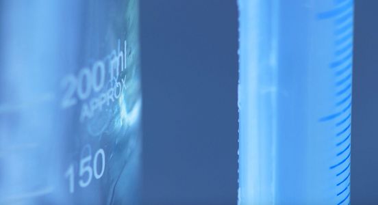 Film-Still: Nahaufnahme einer Art Messbehältnis mit Mililiter-Strichelung. Das Film-Still ist überwiegend in einem warmen Blauton gehalten.