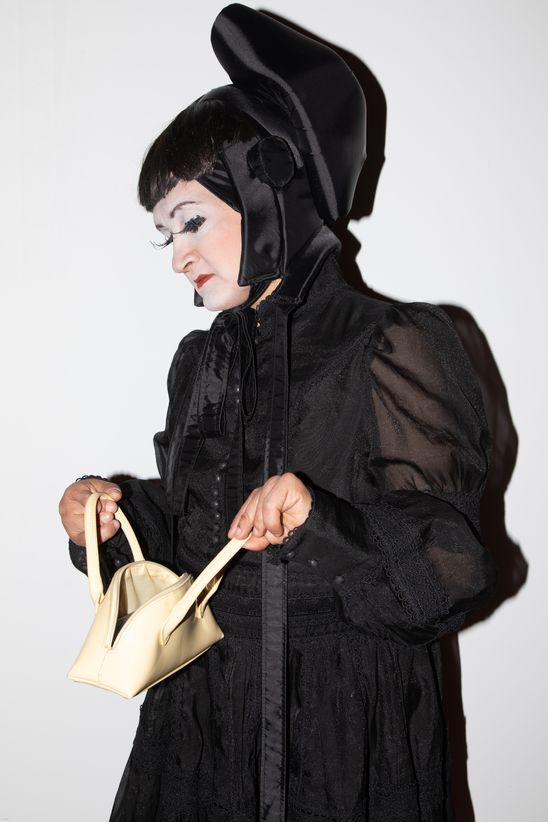 Foto: BayBJane in schwarzem, hochgeschlossenem Kleid mit Hut und cremefarbener kleiner Handtasche.