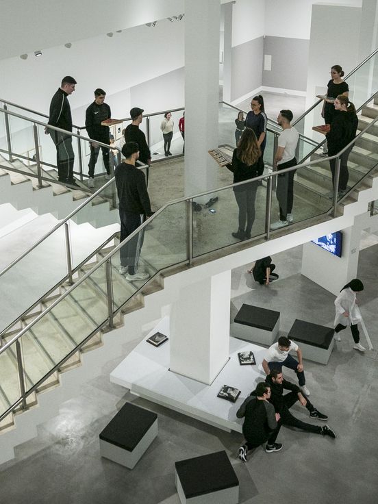 Foto: Viele Jugendliche stehen und sitzen verteilt in einem großen Raum mit zwei Treppen. Zuschauer*innen sehen ihnen bei der Performance zu.