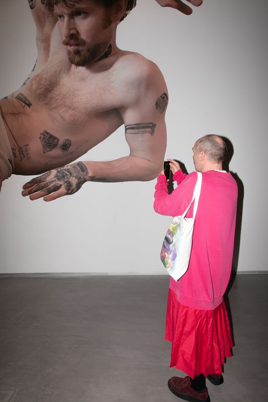 Foto: Eine Person mit langem rotem Rock und leuchtend pinkem Pullover steht vor einer Foto-Tapete von Marc Brandenburg und macht ein Foto.