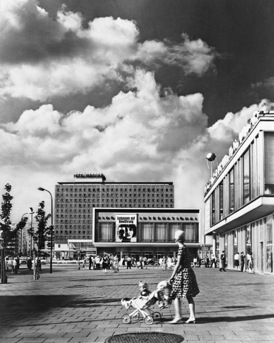 Schwarz-Weiß-Fotografie eines weiten städtischen Platzes mit Passantinnen und Passanten, moderner Architektur und Wolkenformationen am Himmel