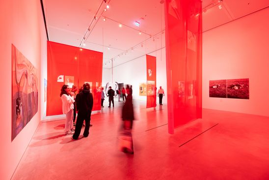 Ausstellungsansicht „Özlem Altın“: Blick in den Ausstellungsraum, der durch rot-orangene Folien, die als Foto-Installationen von der Decke hängen, in farbiges Licht getaucht ist.