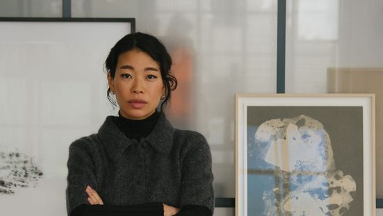 Eine Frau steht mit verschränkten Armen in einem Atelier vor einem Tisch mit Kunstwerken, gerahmten Bildern und Materialien. Sie trägt dunkle Kleidung und blickt direkt in die Kamera.