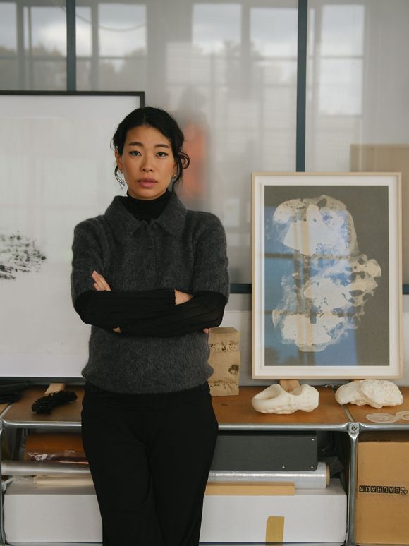 Eine Frau steht mit verschränkten Armen in einem Atelier vor einem Tisch mit Kunstwerken, gerahmten Bildern und Materialien. Sie trägt dunkle Kleidung und blickt direkt in die Kamera.