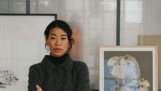 A woman stands with her arms crossed in a studio in front of a table with artworks, framed images, and materials. She is wearing dark clothing and looks directly at the camera.