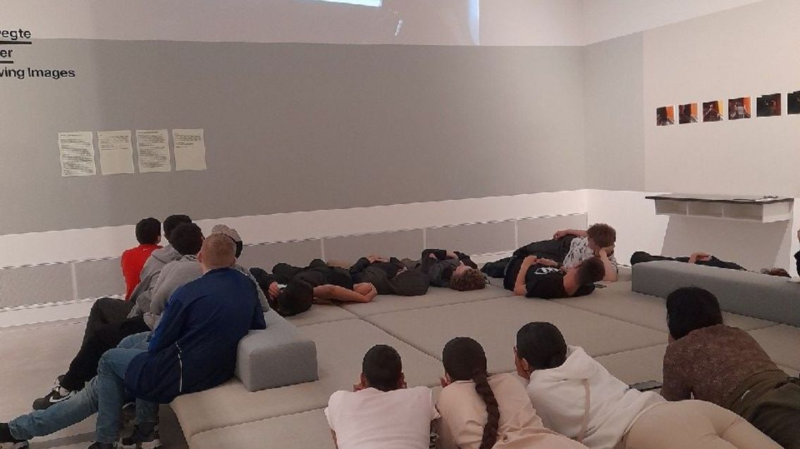 Die Klasse liegt auf dem Couchmöbel im Vermittlungsraum der Berlinischen Galerie und schaut nach oben an die Wand, auf die gerade ihre Videoarbeiten projiziert werden.