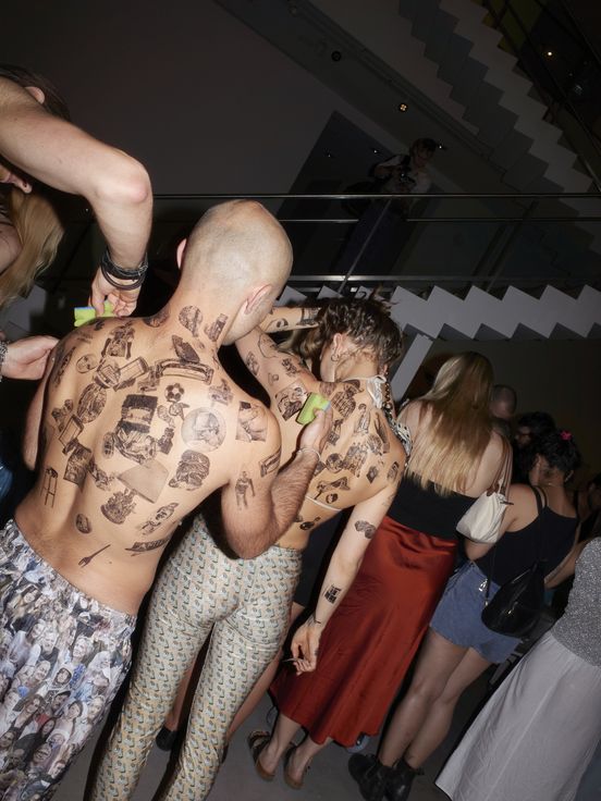 Foto: Mit Blitz aufgenommene Rückenansicht von Personen, die sich gegenseitig den Oberkörper mit temporären Tattoos bekleben.