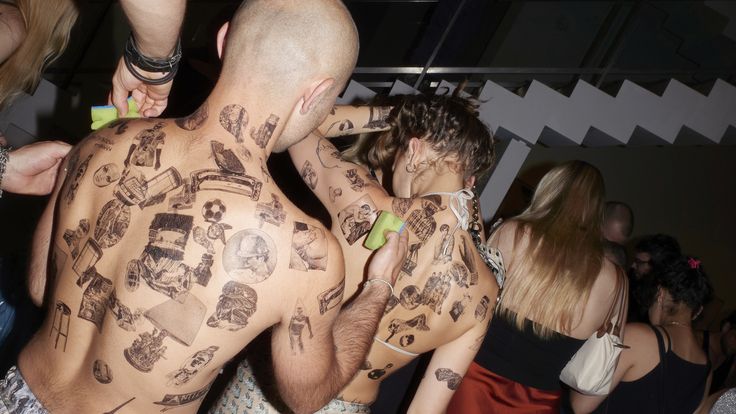 Photo: A flash photograph showing the backs of people applying temporary tattoos to each other’s upper bodies.