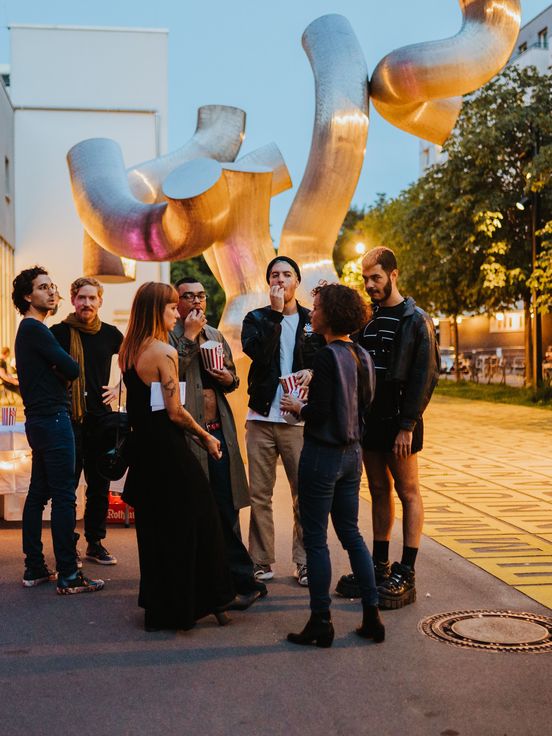 Foto: Besucher*innen stehen draußen rechts neben dem Museum, zum Teil schick gekleidet. Im Hintergrund ist eine silberne Skulptur und das gelbe Buchstabendfeld, der Vorplatz der Berlinischen Galerie, zu sehen.