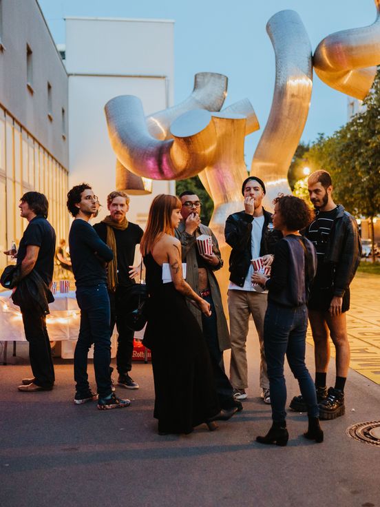 Photo: Visitors stand outside to the right of the museum, some of them smartly dressed. In the background you can see a silver sculpture and the yellow field of letters, the forecourt of the Berlinische Galerie.