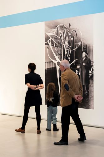 Foto: Drei Personen stehen vor einer schwarz-weiß-Fototapete, die Hans Uhlmann neben einer seiner Skulpturen zeigt.