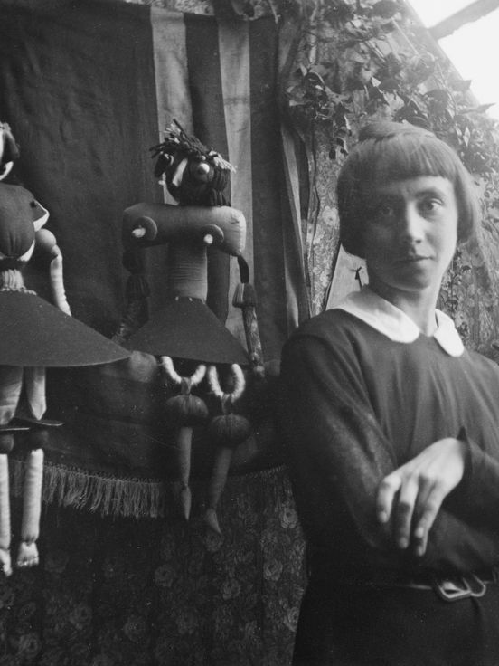 Schwarz-weiß-Fotografie, die Hannah Höch vor einem Fenster und im Hintergrund zwei Puppen zeigt.