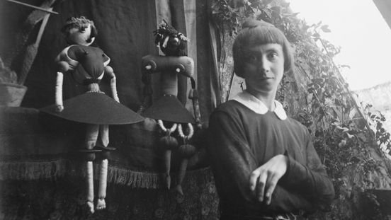 A black-and-white photograph showing Hannah Höch in front of a window, with two dolls in the background.