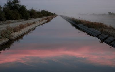 Photo: A view of a long canal in a misty landscape. A pink-coloured sky is reflected on the foreground section of the canal.