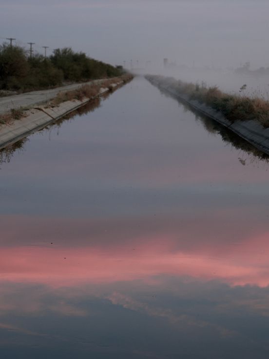 Foto: Blick auf einen langen Kanal in einer nebeligen Landschaft. Auf dem vorderen Teil des Kanals spiegelt sich ein rosa-farbener Himmel.