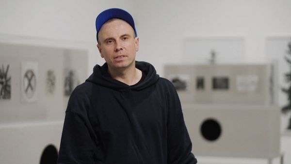 Video still: A person in a dark blue cap and hoodie stands in front of grey glass display cases showing photographs.