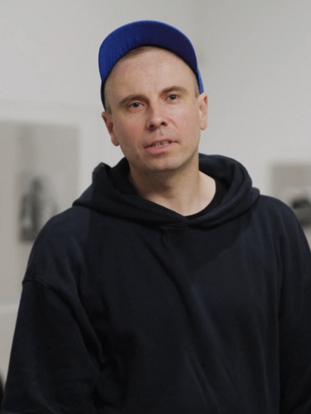 Video still: A person in a dark blue cap and hoodie stands in front of grey glass display cases showing photographs.