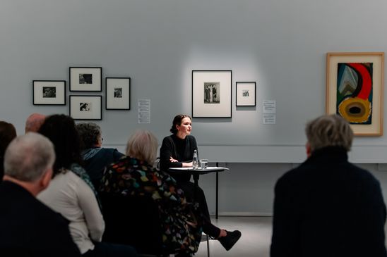 Photo: Melika Foroutan sits at a small table in the exhibition room in front of an audience.