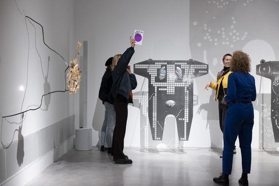 Photo: Four people stand in an exhibition space between works of art by Mariechen Danz and talk. One person holds up a brochure for Berlin Art Week 2024.