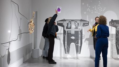 Photo: Four people stand in an exhibition space between works of art by Mariechen Danz and talk. One person holds up a brochure for Berlin Art Week 2024.