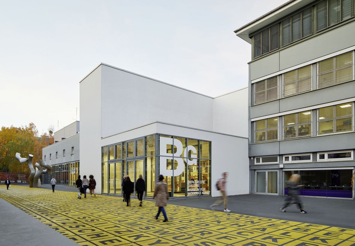 Foto: Gebäude mit Flachdach und Glasfront, auf der das Logo der Berlinischen Galerie abgebildet ist. Davor Menschen auf einem Platz mit Metallskulptur und Bodenkunstwerk.