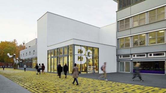 Photo: Building with flat roof and glass front displaying the logo of the Berlinische Galerie. In front of it, people on a square with metal sculpture and floor art.