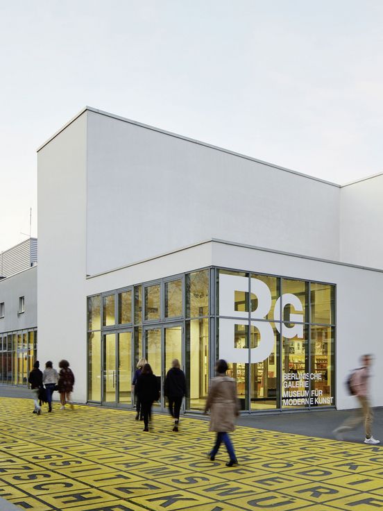 Foto: Gebäude mit Flachdach und Glasfront, auf der das Logo der Berlinischen Galerie abgebildet ist. Davor Menschen auf einem Platz mit Metallskulptur und Bodenkunstwerk.