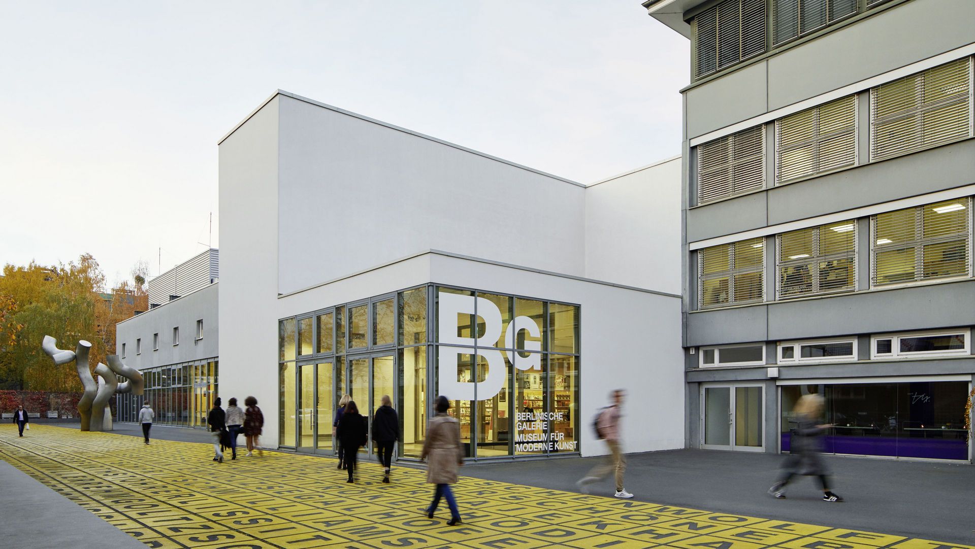 Foto: Gebäude mit Flachdach und Glasfront, auf der das Logo der Berlinischen Galerie abgebildet ist. Davor Menschen auf einem Platz mit Metallskulptur und Bodenkunstwerk.