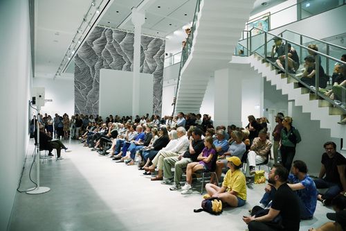 A large group of people is either sitting or standing in the exhibition space, in parts on the staircase crossing the room, while listening to a conversation.