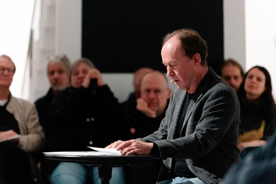 Photo: Ulrich Noethen sits at a small table in the exhibition room in front of an audience.