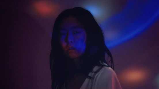 Film still: A person with long dark hair stands in a relatively dark environment, red-blue light is projected onto her and the wall behind her.