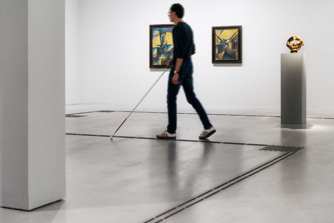 Foto: Besucher mit Blindenlangstock folgt dem Bodenleitsystem im Ausstellungsraum.