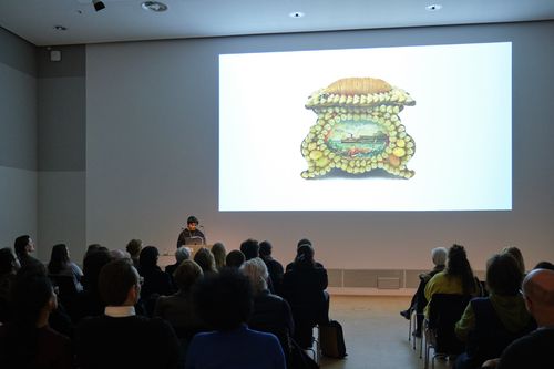 Photo: Monira Al Qadiri stands in front of an audience at a lectern. The background depicts a screened part of the artists presentation.
