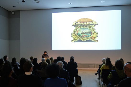 Photo: Monira Al Qadiri stands in front of an audience at a lectern. The background depicts a screened part of the artists presentation.