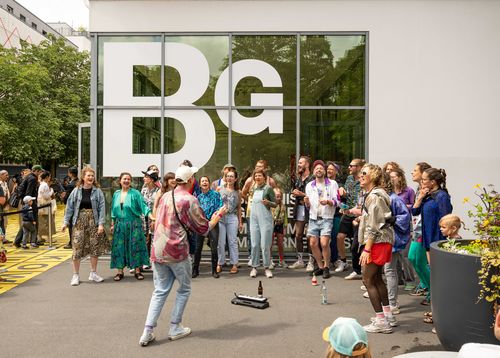 Concert by Projekt Kiezchor Photo: A group of colourfully dressed people stand in a semi-circle in front of the Berlinische Galerie and sing.