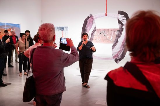 Foto: Die Künstlerin während des Rundgangs durch den Ausstellungsraum. Um sie herum stehen viele Teilnehmende und machen zum Teil Fotos. Im Hintergrund sind einige Arbeiten zu sehen.