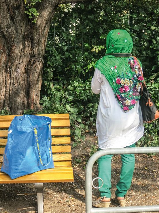 Foto: Eine Frau steht uns mit dem Rücken zugewandt neben einer Parkbank. Eine Kameraschlaufe hängt um ihren Hals und sie blickt auf Sträucher und Bäume.