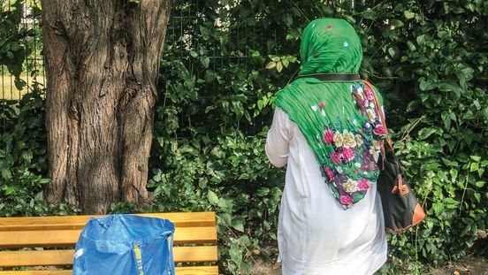 Foto: Eine Frau steht uns mit dem Rücken zugewandt neben einer Parkbank. Eine Kameraschlaufe hängt um ihren Hals und sie blickt auf Sträucher und Bäume.