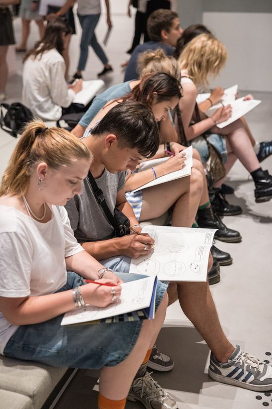 Foto: Junge Menschen sitzen auf einer Sitzplattform und zeichen in Malheften.