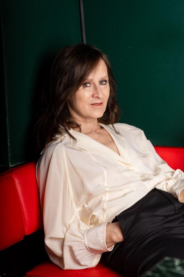 Porträtfoto von Sophie Rois in weißer Bluse auf einer leuchtend roten Bank sitzend.