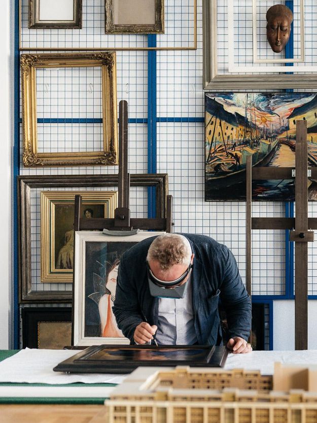 Photo: Person with magnifying glasses looking at a work of art with brush in the hand. Behind on the wall frames and artworks.