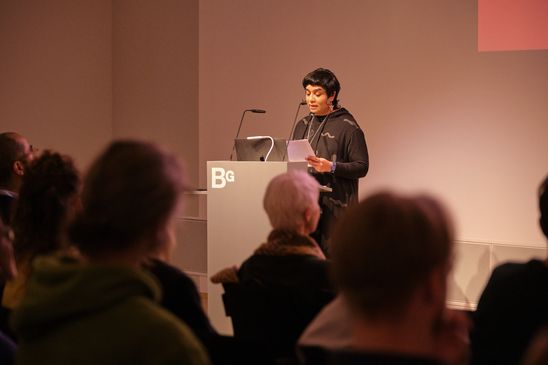 Photo: Monira Al Qadiri stands in front of an audience at a lectern.