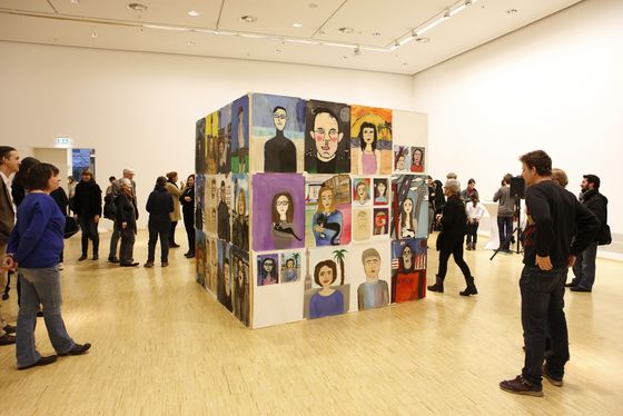 Foto: Viele Menschen stehen um einen großen Würfel im Ausstellungsraum. Auf dem Würfel sind viele von den Schüler*innen gemalte Portraits zu sehen.