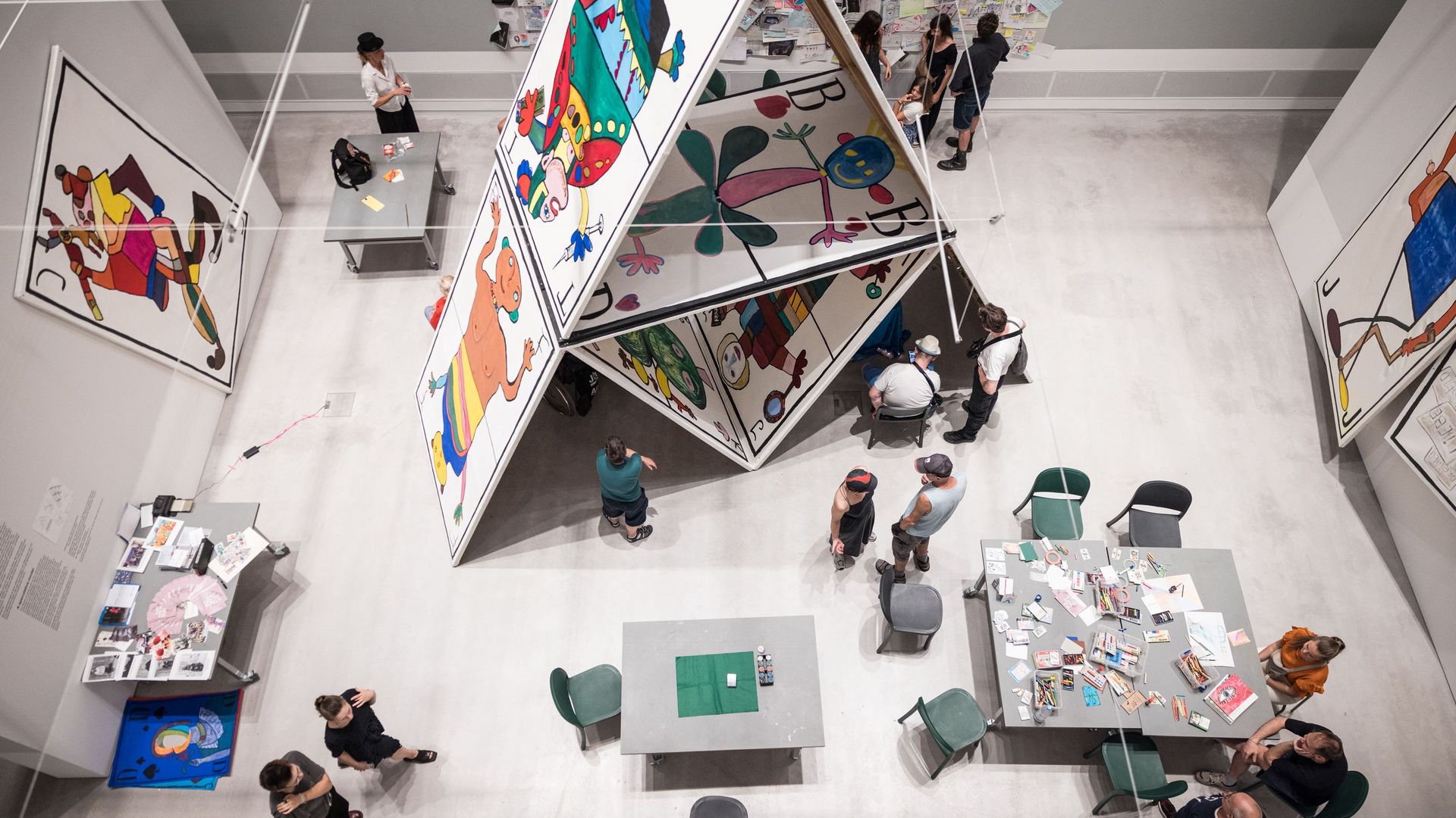 Foto: Blick von oben auf die überlebensgroße Installation „Das Kartenhaus“ der Kunstwerkstatt Kreuzberg der Lebenshilfe Berlin. Neben und unten im Kartenhaus sitzen Menschen an Tischen, reden und basteln.