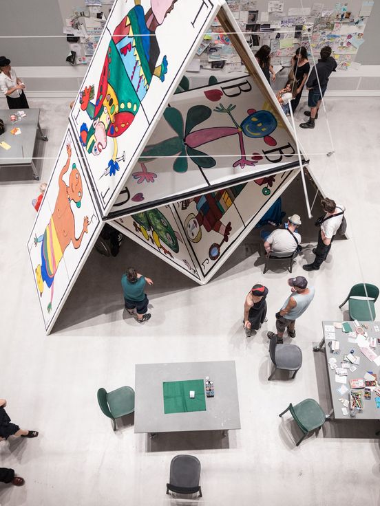 Foto: Blick von oben auf die überlebensgroße Installation „Das Kartenhaus“ der Kunstwerkstatt Kreuzberg der Lebenshilfe Berlin. Neben und unten im Kartenhaus sitzen Menschen an Tischen, reden und basteln.