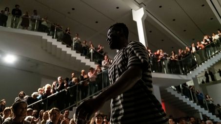 Foto: Meier Eden bei seiner Performance. Im Hintergrund sind viele Zuschauer*innen auf Stühlen, stehend und auf zwei großen Treppen stehend zu sehen.