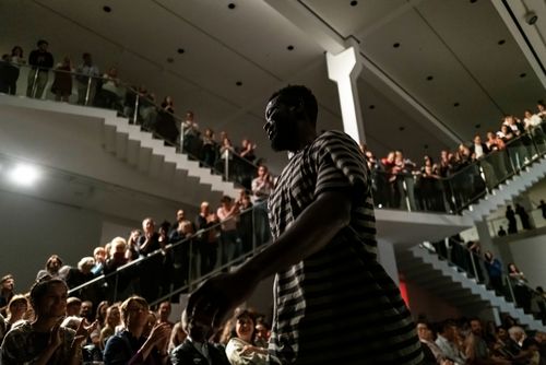 Foto: Meier Eden bei seiner Performance. Im Hintergrund sind viele Zuschauer*innen auf Stühlen, stehend und auf zwei großen Treppen stehend zu sehen.