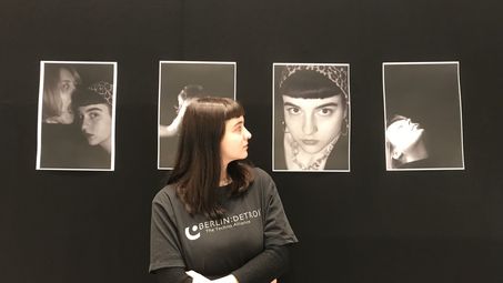 Foto: Eine junge Frau steht vor vier Fotoporträts. Sie blickt auf die Fotografie, welche sie selbst zeigt.